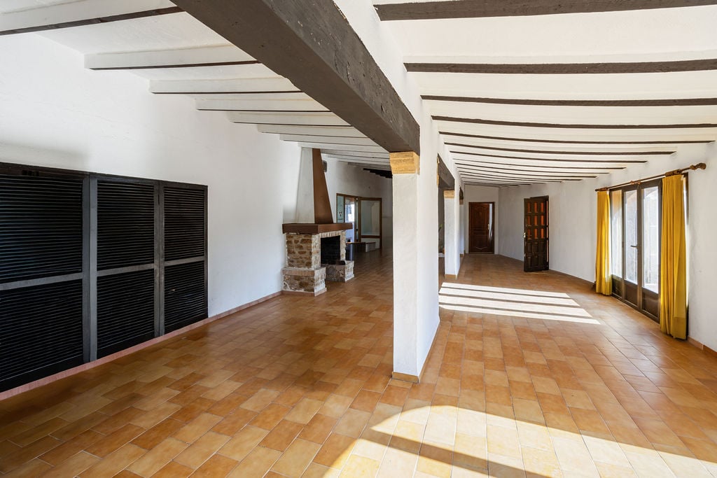 Ruime open kamer met authentieke houten plafondbalken, terracotta tegelvloer en een rustieke stenen open haard. Grote ramen zorgen voor een natuurlijke lichtinval.