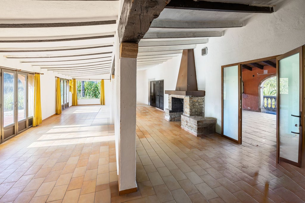 Ruime rustieke woonkamer met terracotta tegelvloer, houten plafondbalken en een stenen open haard. Grote raampartijen zorgen voor een uitstekende natuurlijke lichtinval.