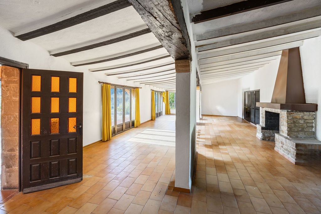 Ruime rustieke zaal met zichtbare donkere houten balken, traditionele terracotta vloer en een natuurstenen open haard met metalen kap.