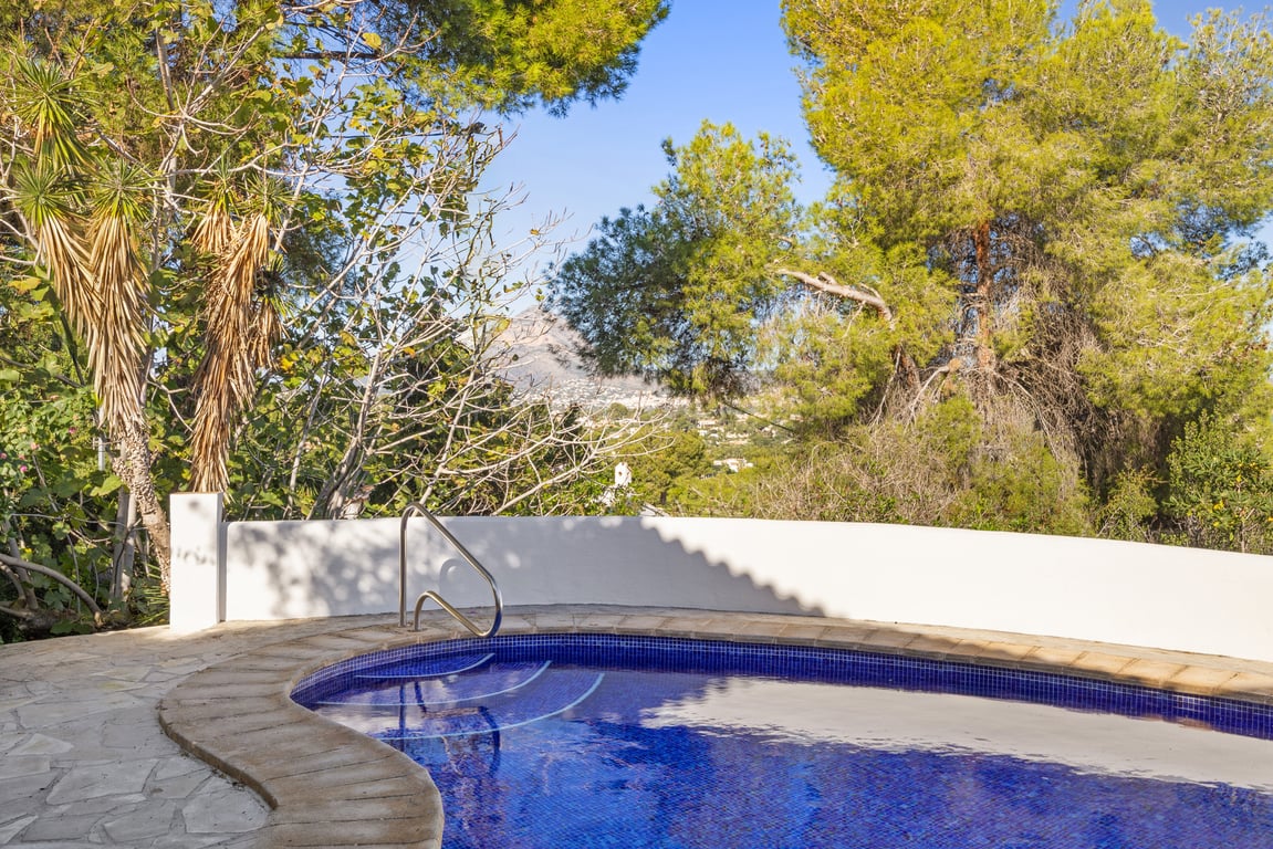 Blauw betegeld zwembad met metalen trapleuning, omgeven door een natuurstenen terras en mediterrane begroeiing met uitzicht op de Montgó-berg.