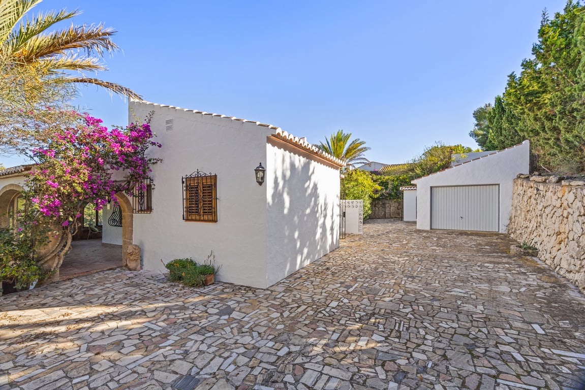 Buiteningang met onregelmatige natuurstenen bestrating, vrijstaande garage en witte gevel versierd met roze bougainville en traditionele Tosca stenen bogen.