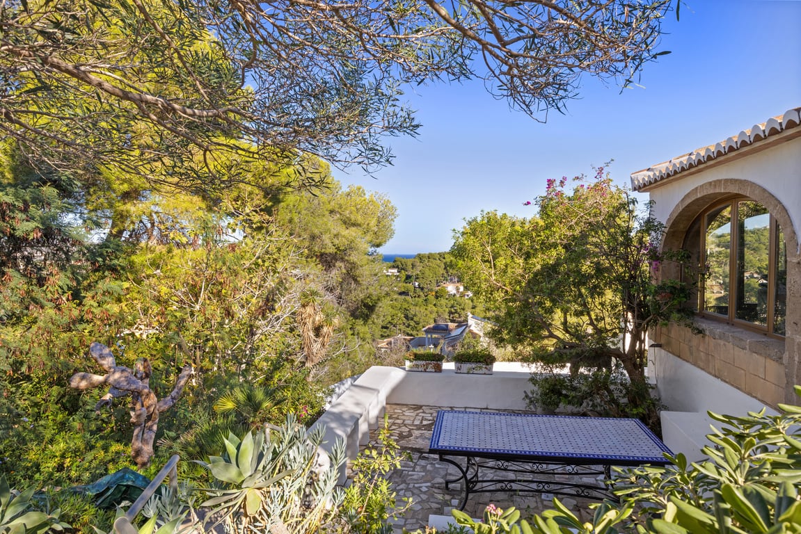Buitenterras met natuurstenen bestrating en een mozaïektafel, omgeven door mediterrane beplanting met uitzicht op zee door de pijnbomen en traditionele architectuur.