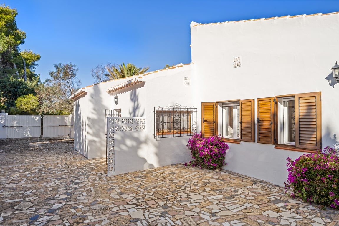 Exterieur van de villa met witte gevel, houten luiken, smeedijzeren roosters en onregelmatige natuurstenen bestrating met bougainvillea.