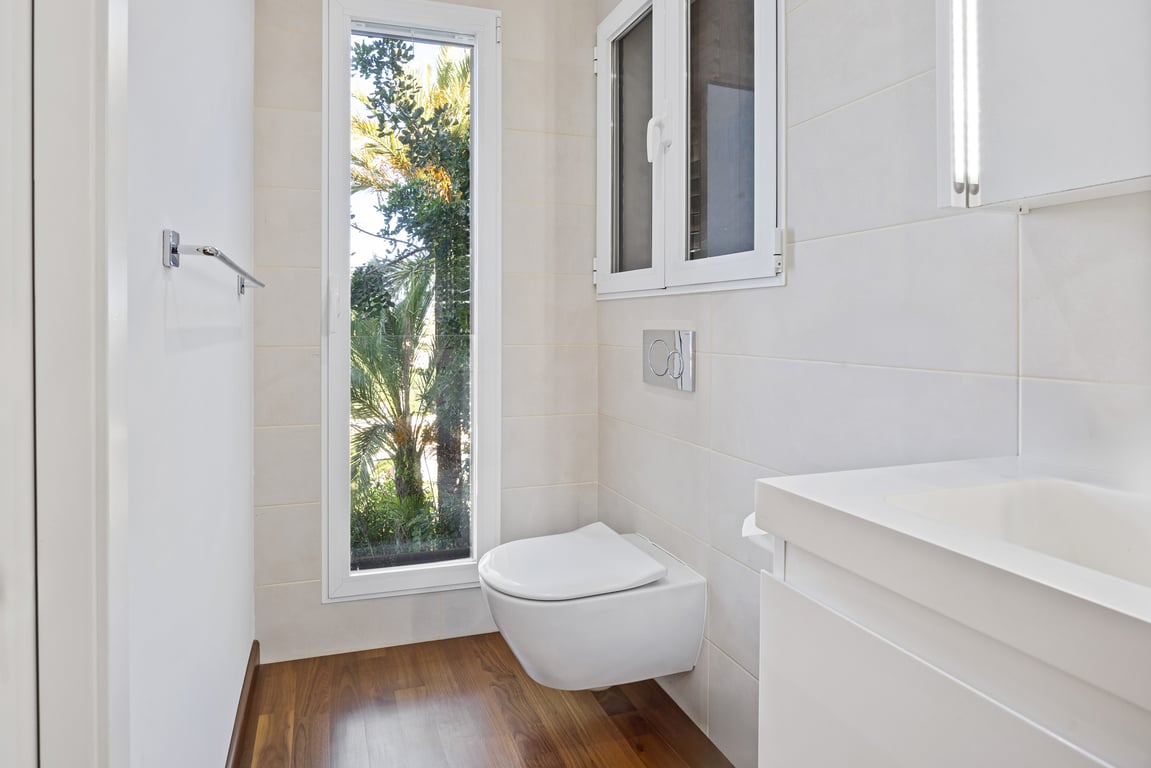 Gerenoveerde badkamer met hangend toilet, houten vloer en een verticaal raam met uitzicht op de omliggende tuin.