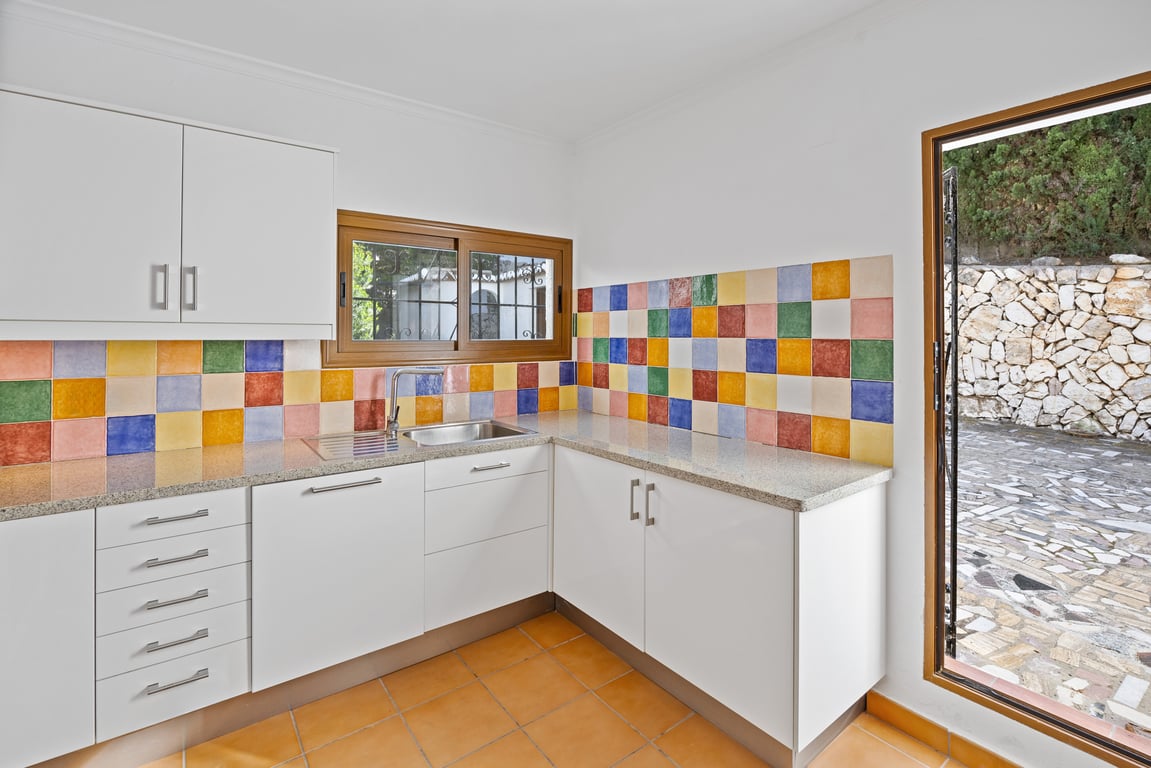 Keuken met witte kasten, een granieten aanrechtblad en een opvallende kleurrijke achterwand. Voorzien van een terracotta vloer en een directe deur naar het buitengedeelte.