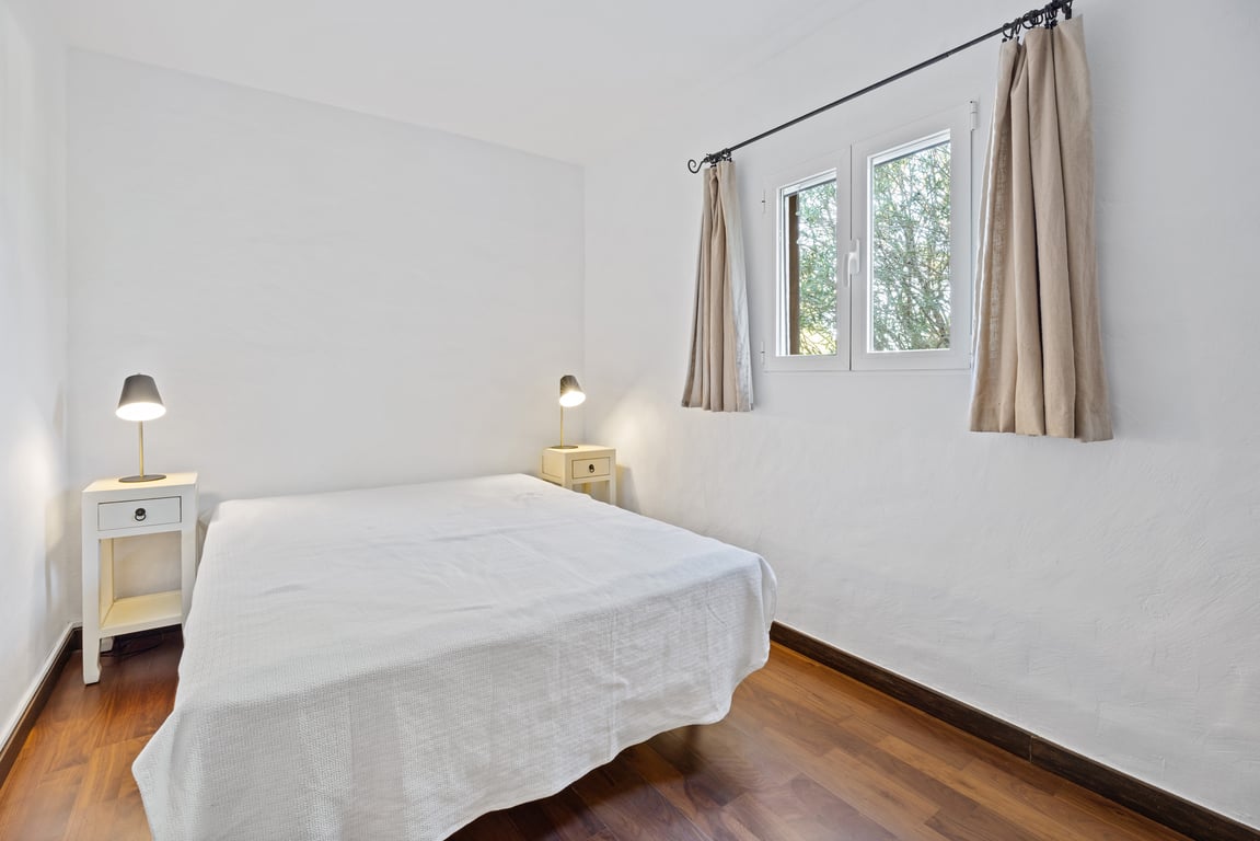 Lichte slaapkamer met houten vloer, witte muren en een raam met dubbel glas dat uitkijkt op het groen. Voorzien van nachtkastjes en leeslampen.