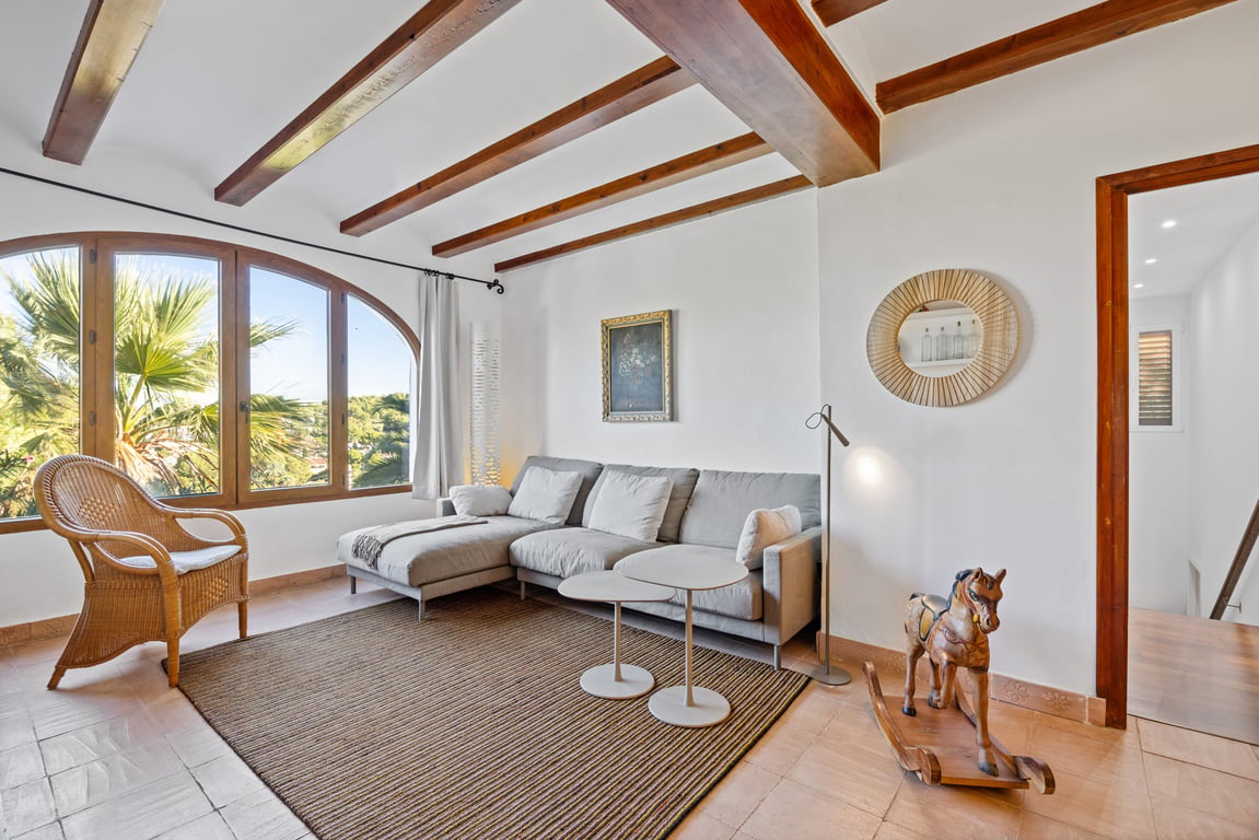 Lichte woonkamer met houten plafondbalken, ambachtelijke tegelvloer en een groot boogvenster met uitzicht op de mediterrane tuin.