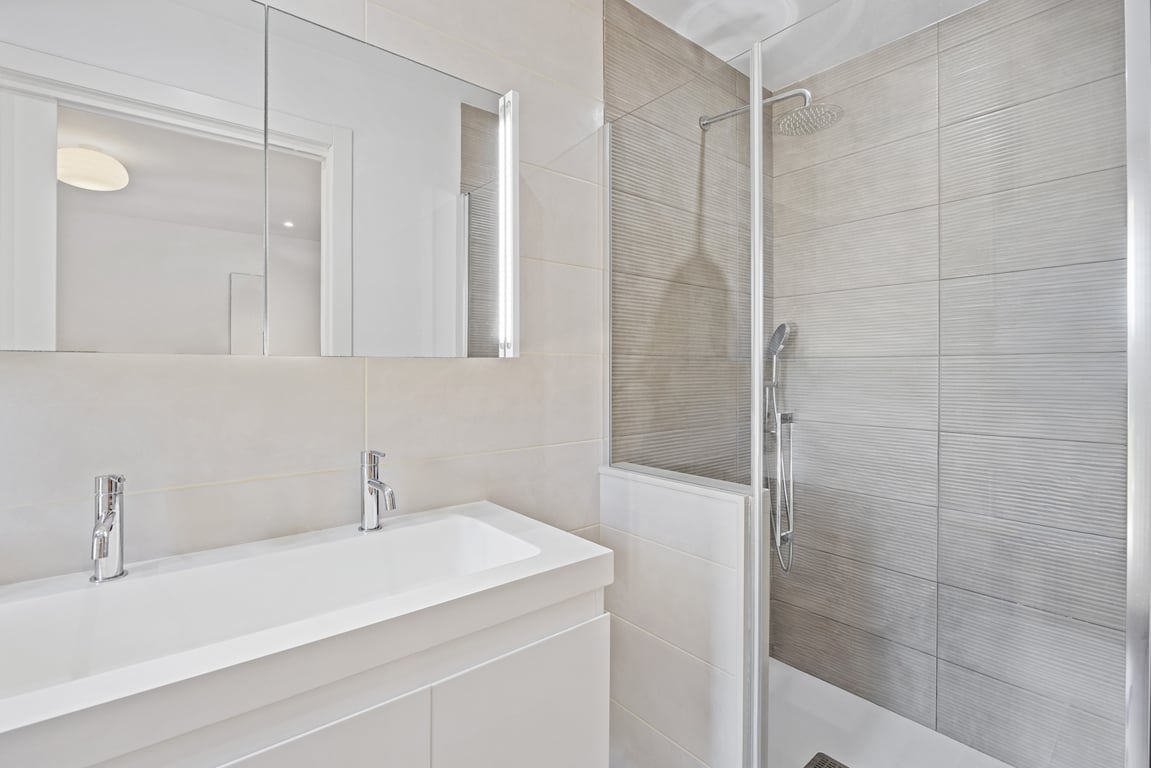 Modern gerenoveerde badkamer met dubbele kraan, witte wastafelkast en inloopdouche met getextureerde grijze tegels en glazen wand.