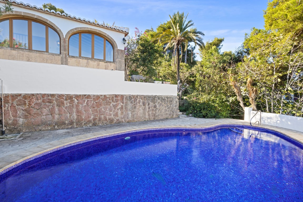 Privézwembad met blauwe mozaïektegels, omgeven door een rustieke stenen muur en terras, met de naya met bogen en mediterrane beplanting op de achtergrond.