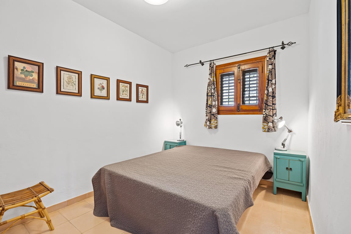 Slaapkamer met tweepersoonsbed, turquoise nachtkastjes en houten raam met traditionele luiken. Witte muren versierd met een reeks botanische prenten.