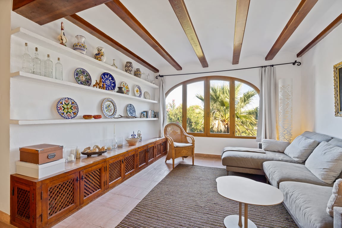Woonkamer met houten balkenplafond, ingebouwde schappen met keramiek en een groot boograam met uitzicht op de mediterrane tuin.