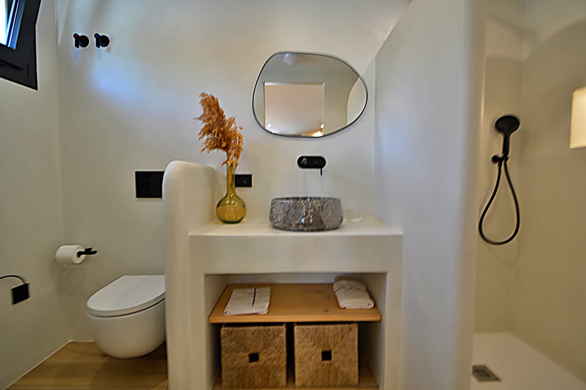 Badkamer in Ibiza-stijl met muren van microcement, een stenen waskom en matzwarte kranen. Voorzien van een organisch gevormde spiegel, inloopdouche en ingebouwde opbergruimte.
