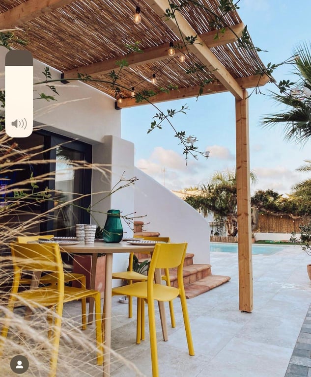 Buiten-eethoek onder een houten pergola met rieten dak en sfeerverlichting. Gele stoelen op een licht stenen terras grenzend aan het zwembad.