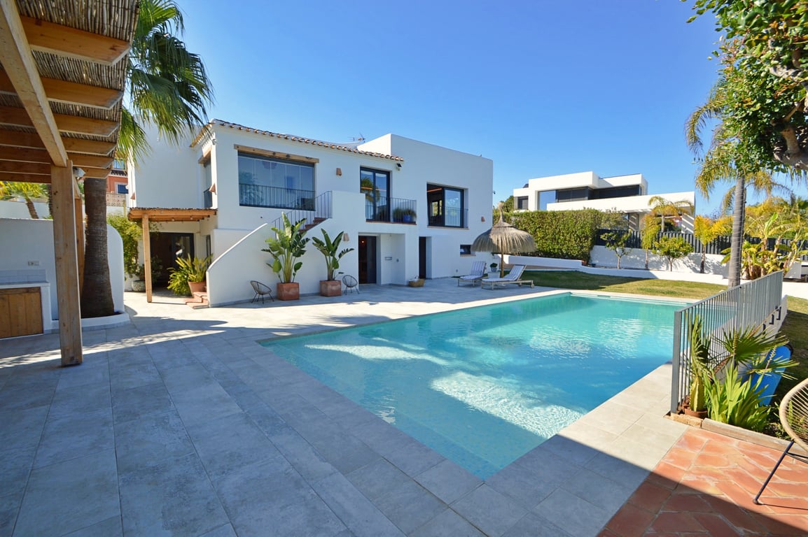Buitenaanzicht van de villa met groot zwembad, licht stenen terras, houten pergola en witte gevel in Ibiza-stijl met een glazen naya op de bovenverdieping.