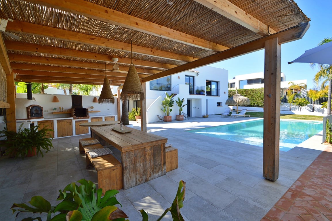 Buitenkeuken onder een houten pergola met pizza-oven, rustieke eethoek en uitzicht op het zoutwaterzwembad en de witte villa in Ibiza-stijl.