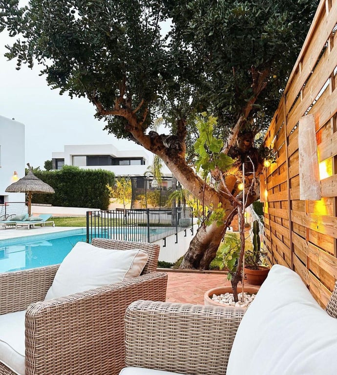Buitenlounge met rotan meubilair op een terracotta terras, voorzien van sfeerverlichting aan de houten schutting nabij het zwembad en een karakteristieke boom.