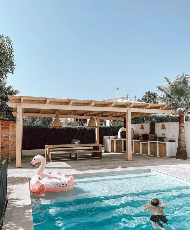 Buitenruimte met eethoek en zomerkeuken voorzien van pizzaoven en barbecue, overdekt door een houten pergola bij het zoutwaterzwembad.
