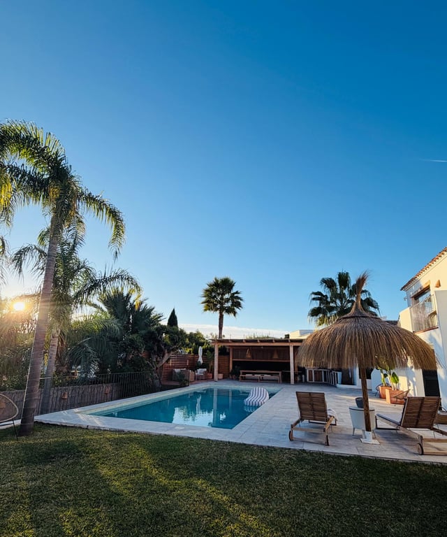 Buitenzwembad met stenen terras, rieten parasol en houten ligbedden. Op de achtergrond een overdekte buitenkeuken en eethoek omgeven door mediterrane beplanting en palmbomen.