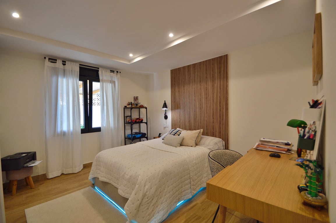 Eigentijdse slaapkamer met houten latten hoofdbord, bureau en bed met blauwe LED-verlichting. Voorzien van een houtlook vloer en verlaagd plafond met inbouwspots.