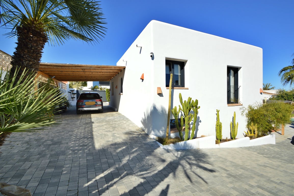 Exterieur in Ibiza-stijl met houten carport, geplaveide oprit en mediterrane tuin met cactussen en palmbomen tegen een strakblauwe lucht.