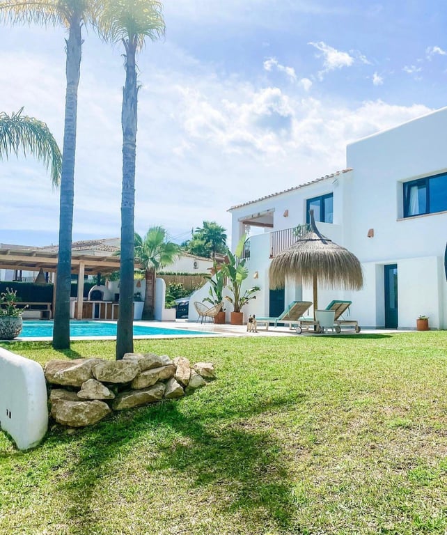 Exterieur in Ibizastijl met gazon, zwembad en rieten parasol. Op de achtergrond is de buitenkeuken met een traditionele pizza-oven te zien.