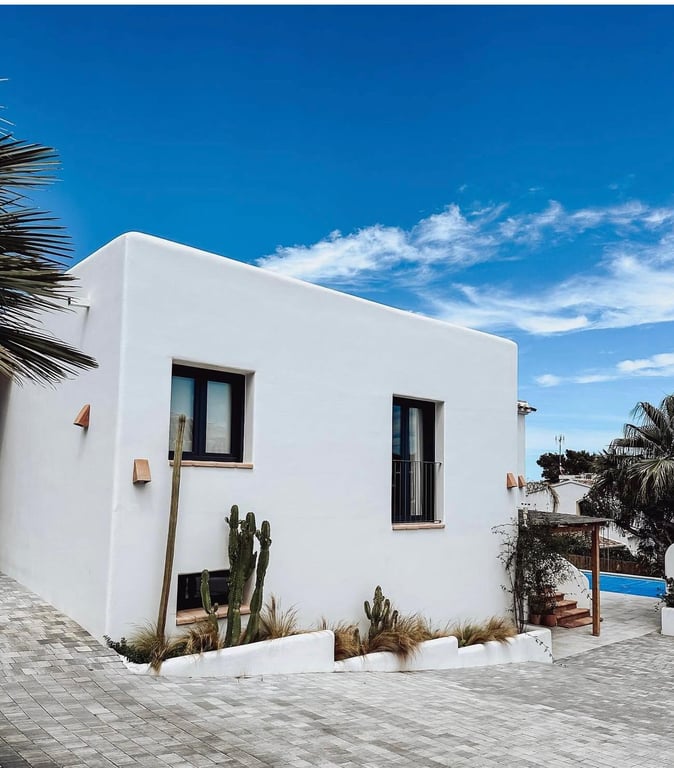 Exterieur in Ibizastijl met gladde witte muren, donkere raamkozijnen en een minimalistische cactustuin langs de oprit van grijze kasseien.