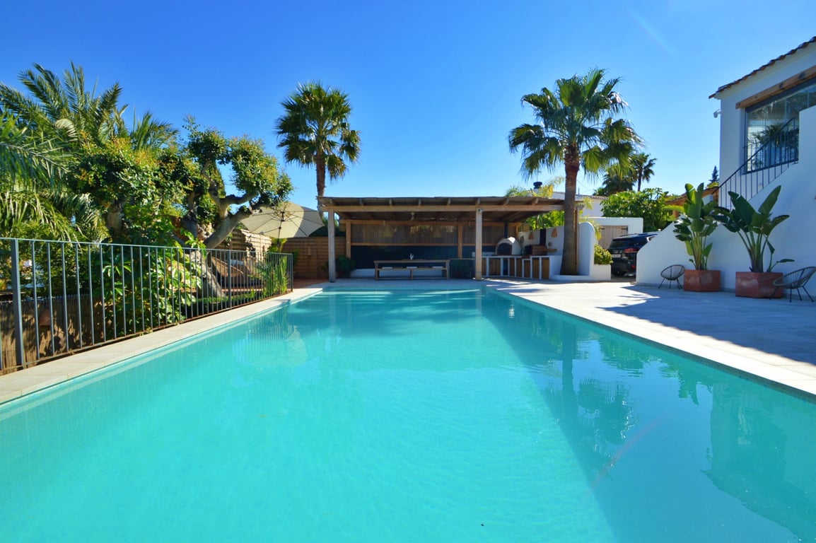 Groot rechthoekig zwembad met ruim zonneterras, voorzien van een overdekte houten pergola voor buiten dineren en een mediterrane tuin met palmbomen.