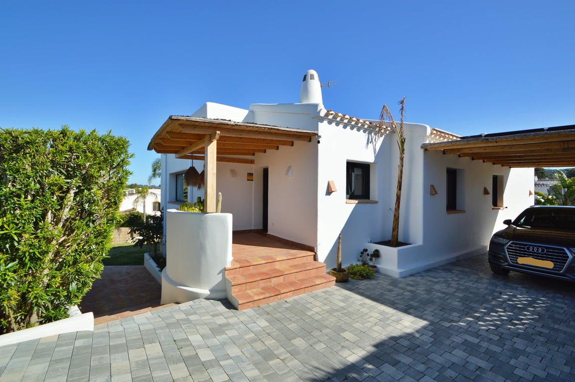 Hoofdingang in Ibiza-stijl met houten pergola en rieten dak, terracotta trappen en een geplaveide oprit die leidt naar een overdekte carport.