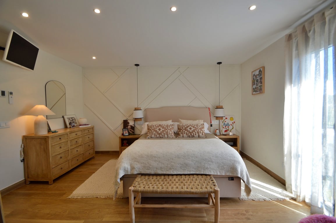 Hoofdslaapkamer met geometrische wandpanelen, lichte houten vloer en inbouwspots. Voorzien van een houten ladekast, hanglampen en een gevlochten bankje bij het voeteneind.