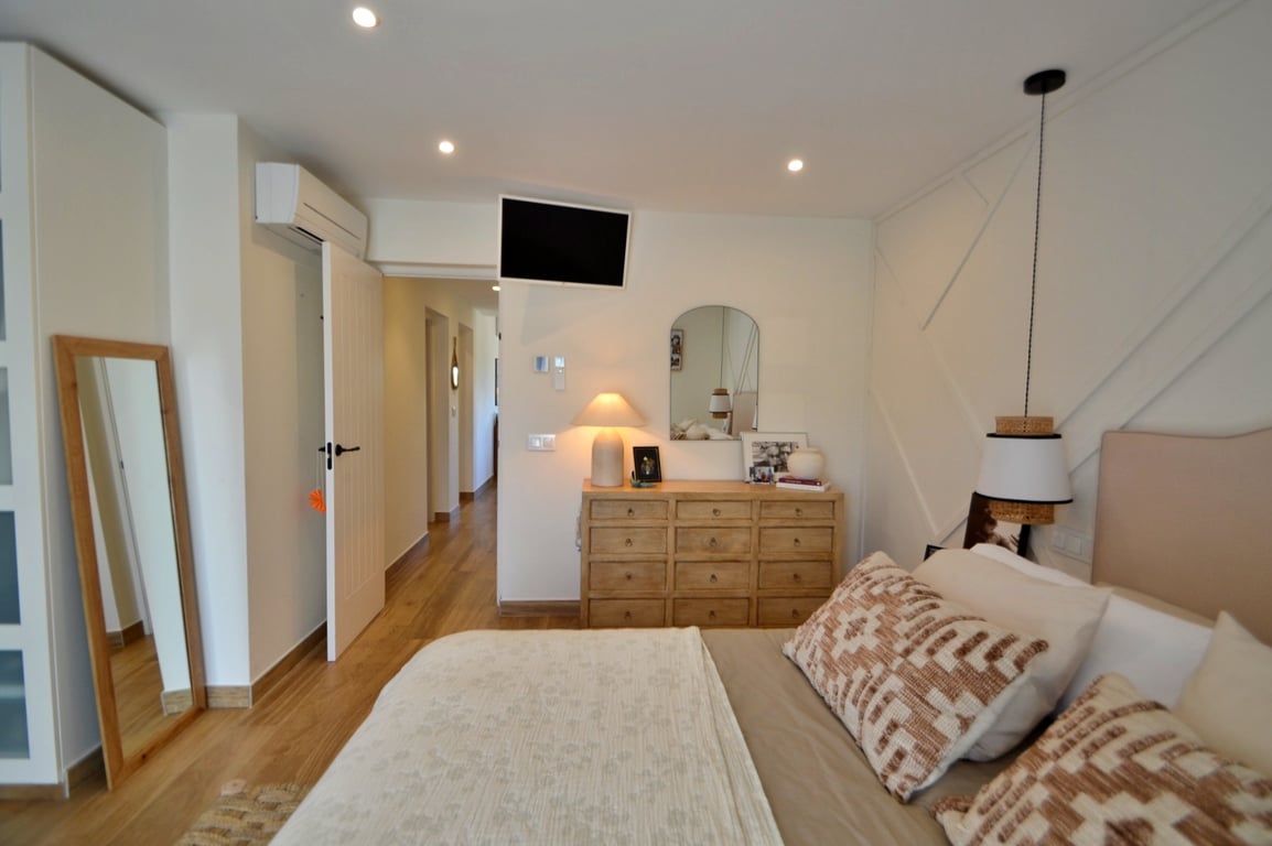 Lichte slaapkamer met houtlook vloer, geometrische wandpanelen, individuele airconditioning en natuurlijke houten meubels zoals een ladekast en passpiegel.