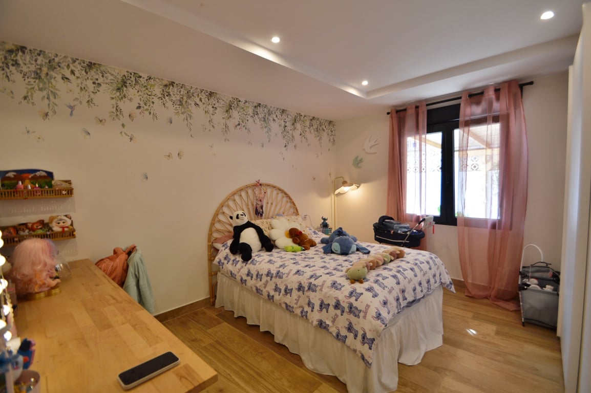 Lichte slaapkamer met rotan hoofdbord, botanische muurschildering en houten vloer. Voorzien van inbouwspots en natuurlijke lichtinval via het grote raam.