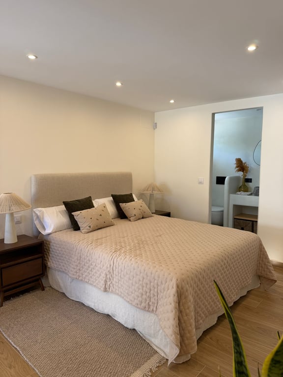 Lichte slaapkamer met tweepersoonsbed, gestoffeerd hoofdbord en houten nachtkastjes. Zicht op de aangrenzende moderne badkamer en keramische vloer met houtlook.