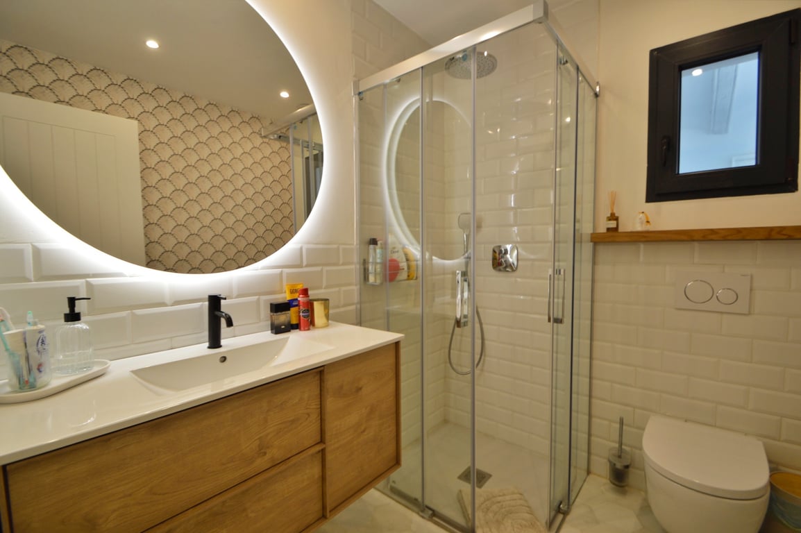 Moderne badkamer met een verlichte ronde spiegel, houten meubel en een glazen douchecabine met witte metrotegels.