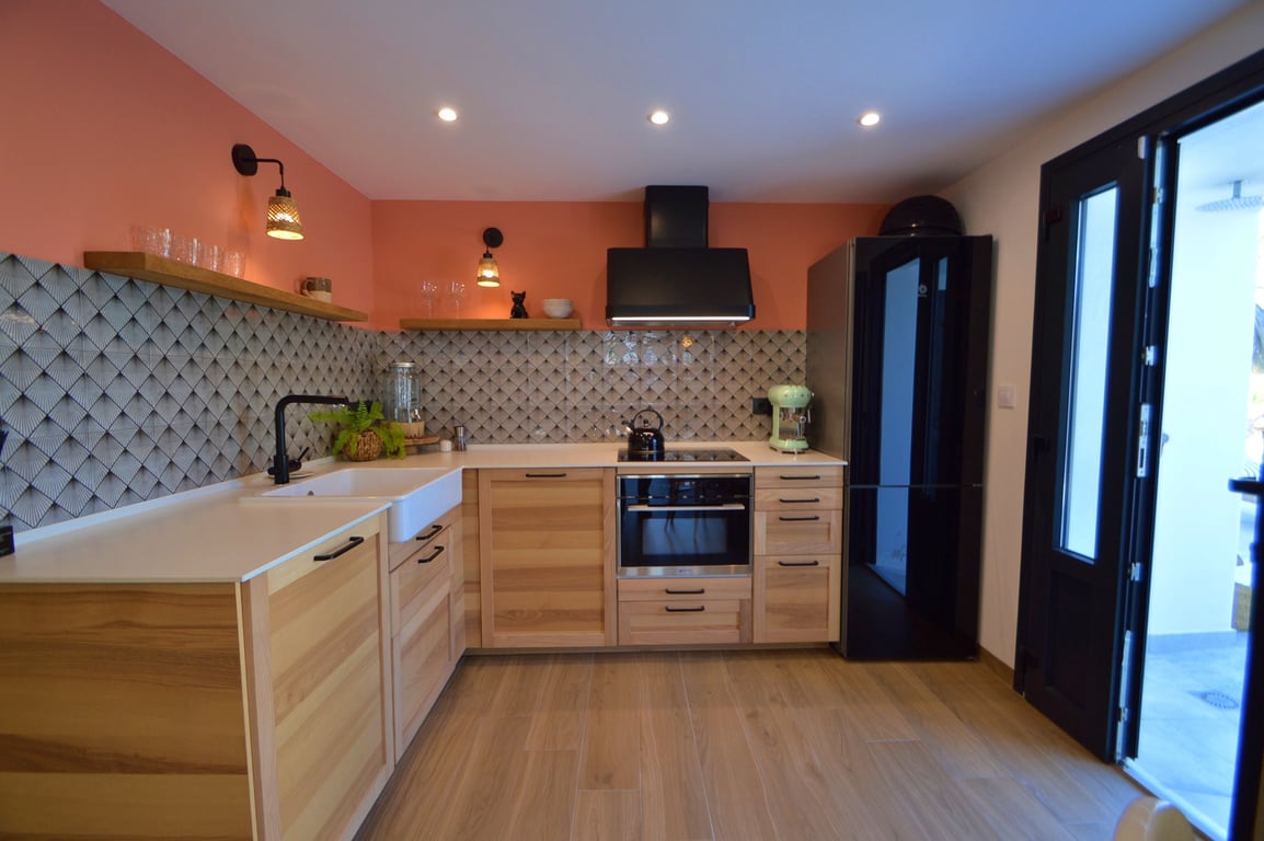 Moderne keuken met lichte houten kasten, een witte landelijke spoelbak en geometrische tegels. Zwarte apparatuur contrasteert met de terracotta accentmuur en houten wandplanken.
