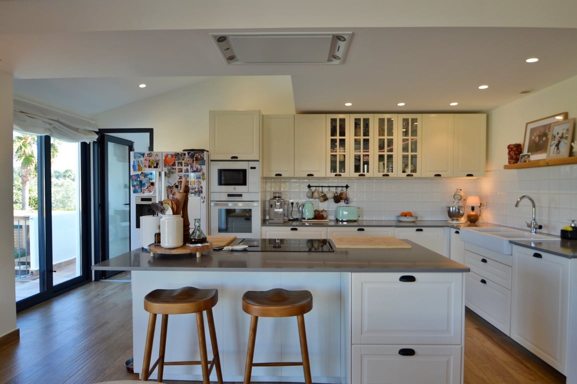 Moderne open keuken met kookeiland, houten krukken en witte kasten. Voorzien van inbouwapparatuur en grote glazen deuren die leiden naar het terras.