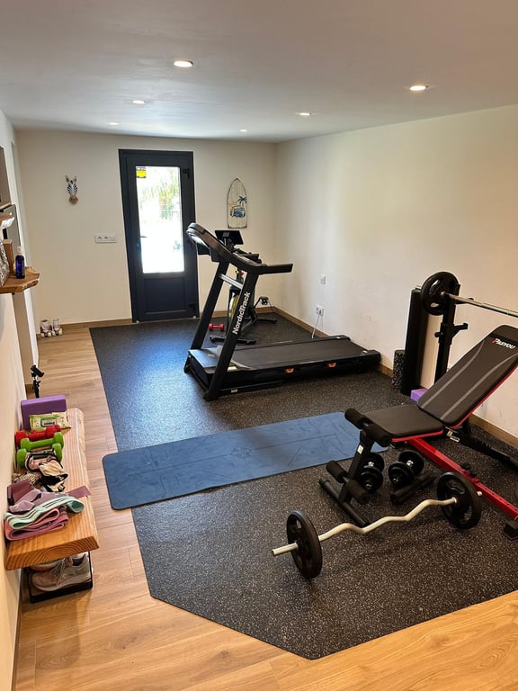 Privé-fitnessruimte met loopband, gewichten en yogamat, voorzien van een houtlook vloer, inbouwspots en directe toegang naar buiten.