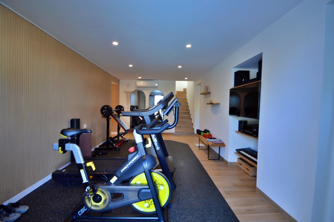 Privé-fitnessruimte met spinningfiets, loopband en gewichten, voorzien van een houten wandpaneel, inbouwspots en een geïntegreerde multimedia-hoek.