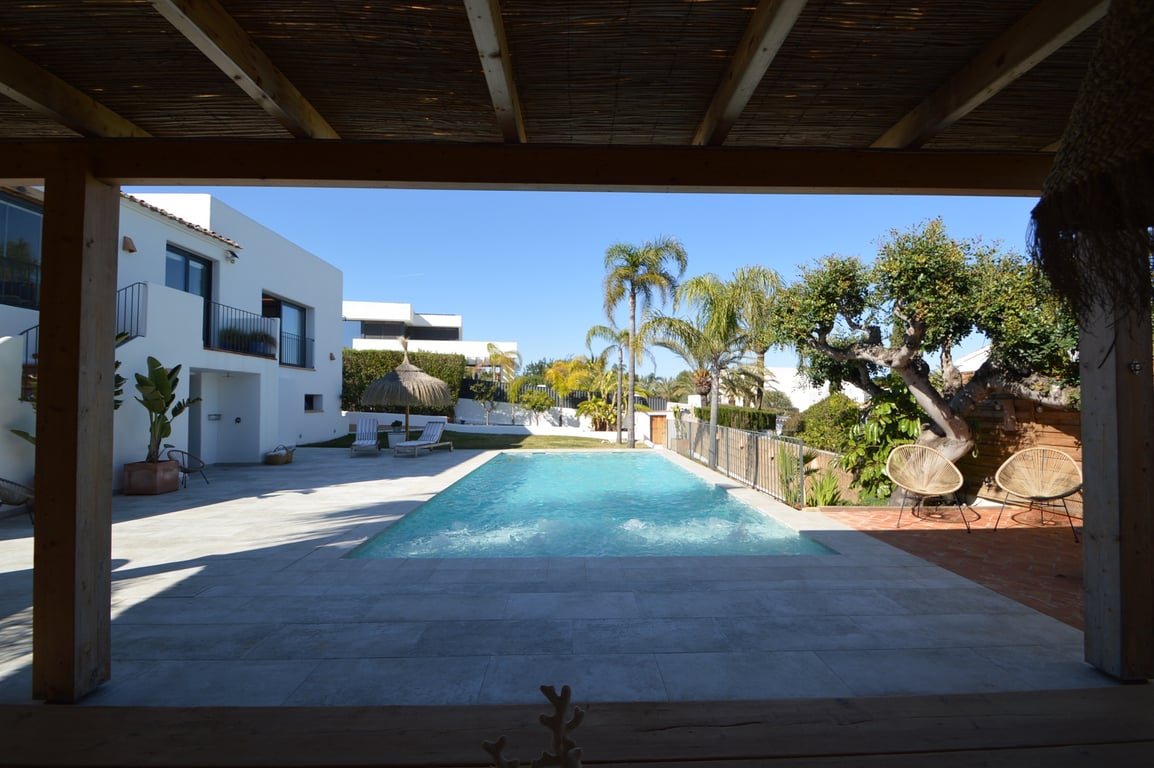 Uitzicht vanaf de houten pergola op het zoutwaterzwembad, omgeven door lichtgrijze terrastegels, palmbomen en de witte gevel in Ibiza-stijl.