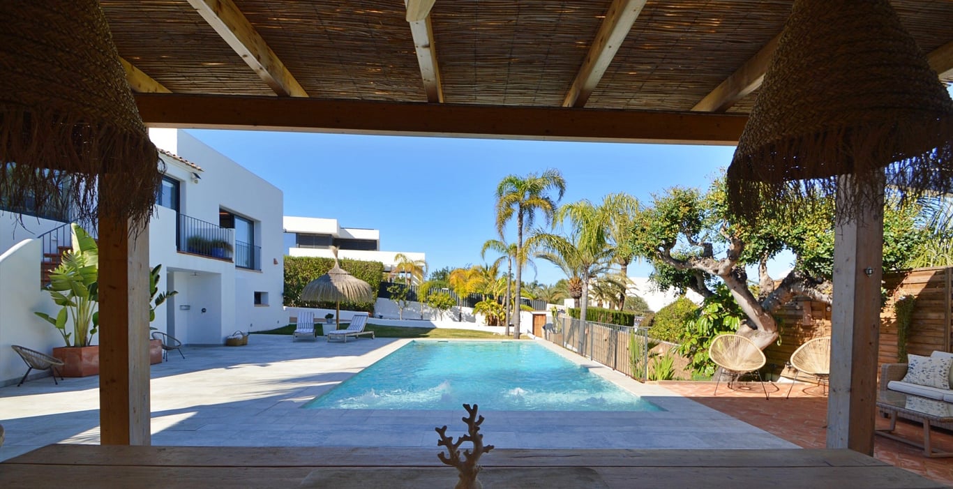 Uitzicht vanaf het schaduwrijke terras met rieten dak op het zoutwaterzwembad en de mediterrane tuin met palmbomen.