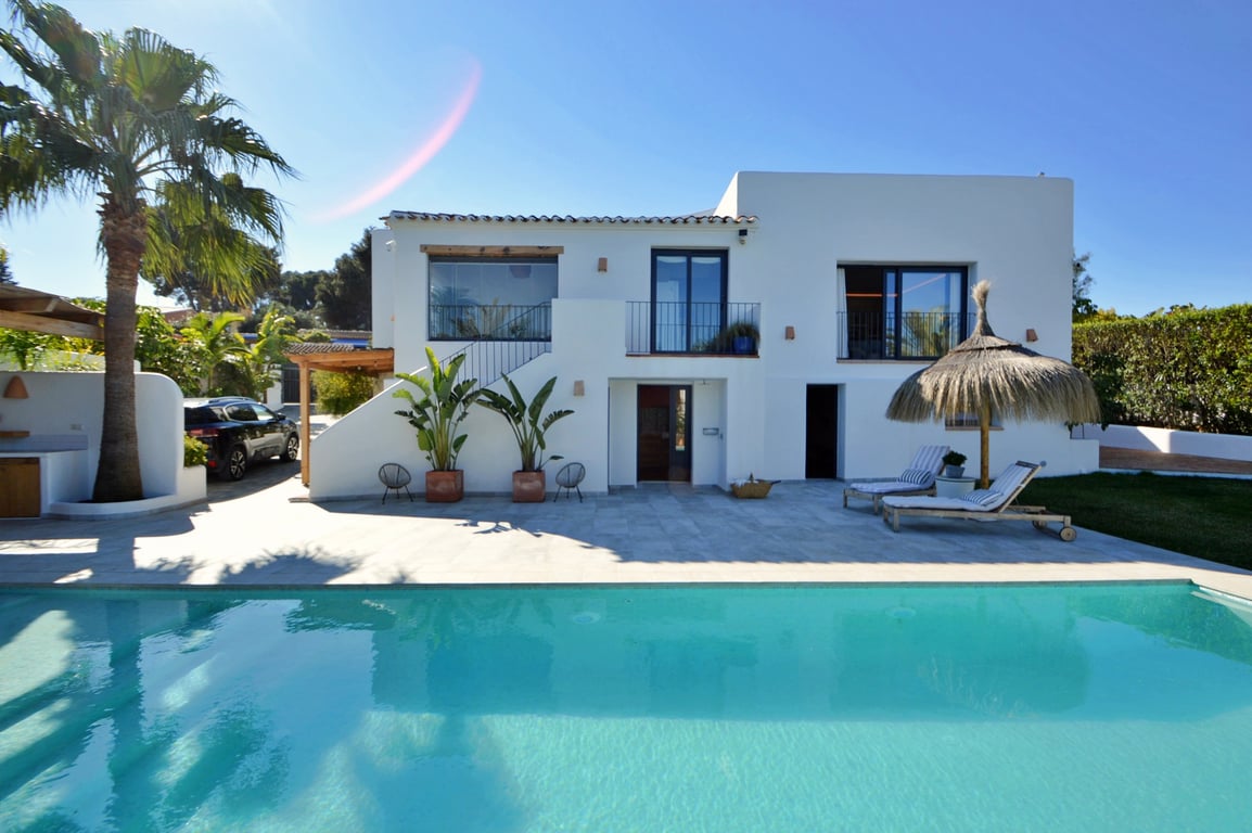 Witte gevel in Ibiza-stijl aan een groot zoutwaterzwembad, met een stenen terras, palmbomen en een loungegedeelte met een rieten parasol.