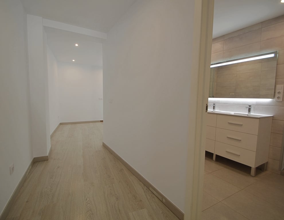 Gang met lichte houten vloer die leidt naar een moderne badkamer met dubbele wastafel, verlichte spiegel en beige wandtegels.