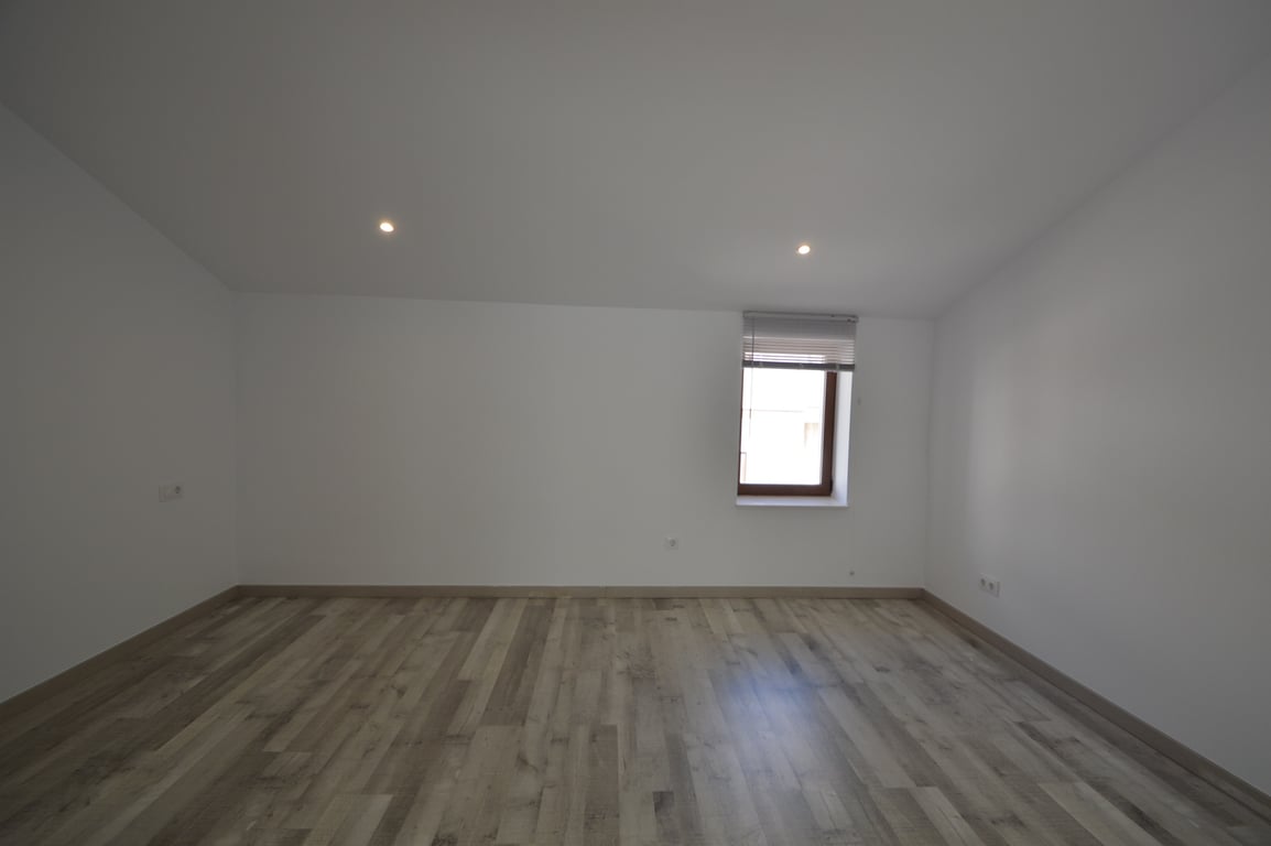 Gerenoveerde kamer met lichte laminaatvloer, gladde witte muren, inbouwspots in het plafond en een raam met jaloezieën.
