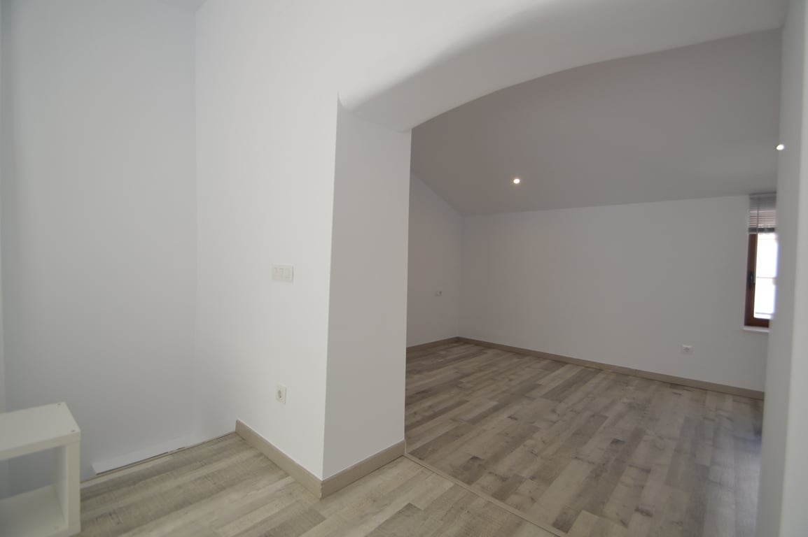Gerenoveerde kamer met lichte laminaatvloer, witte muren en inbouwspots. De ruimte heeft een brede doorgang en een raam met een donker kozijn.