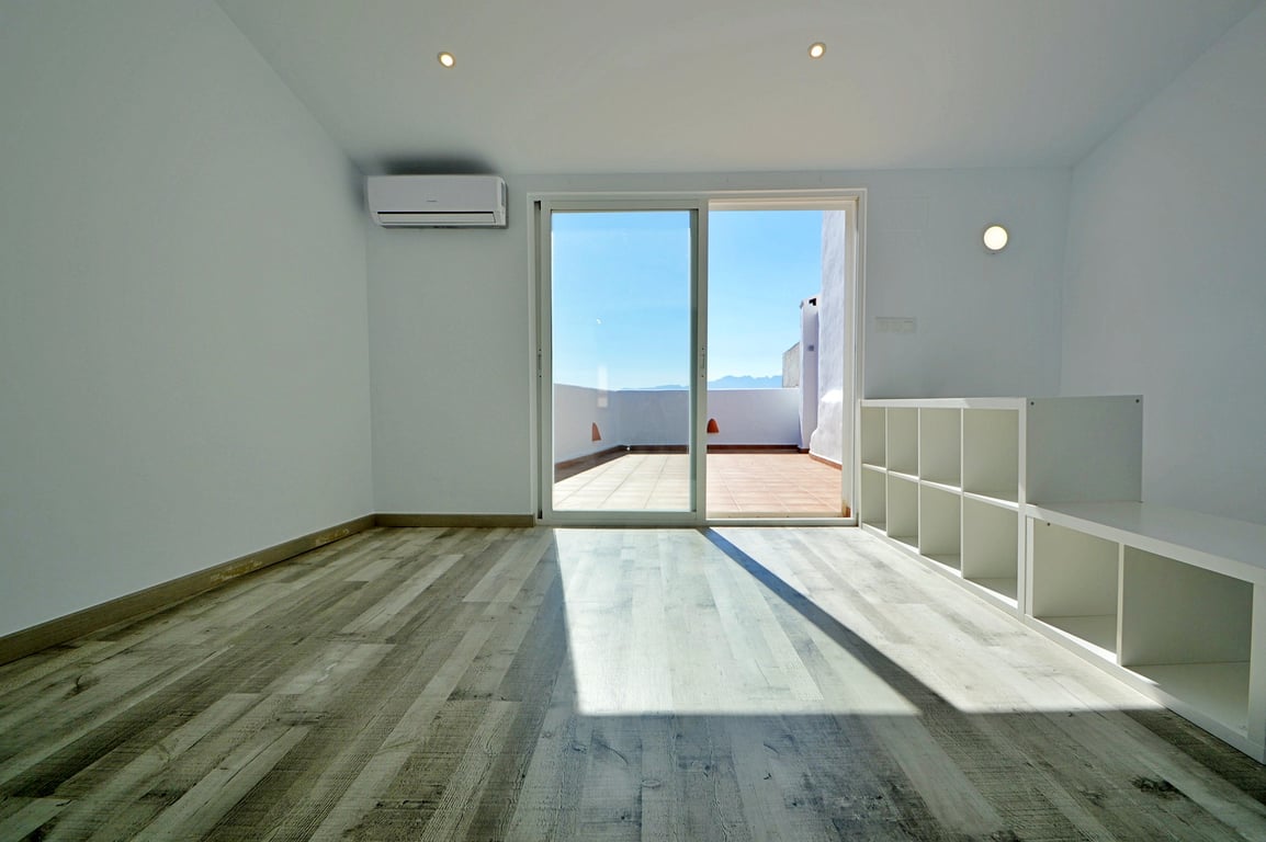 Lichte kamer met laminaatvloer, airconditioning en een glazen schuifpui die toegang geeft tot het ruime dakterras met uitzicht.