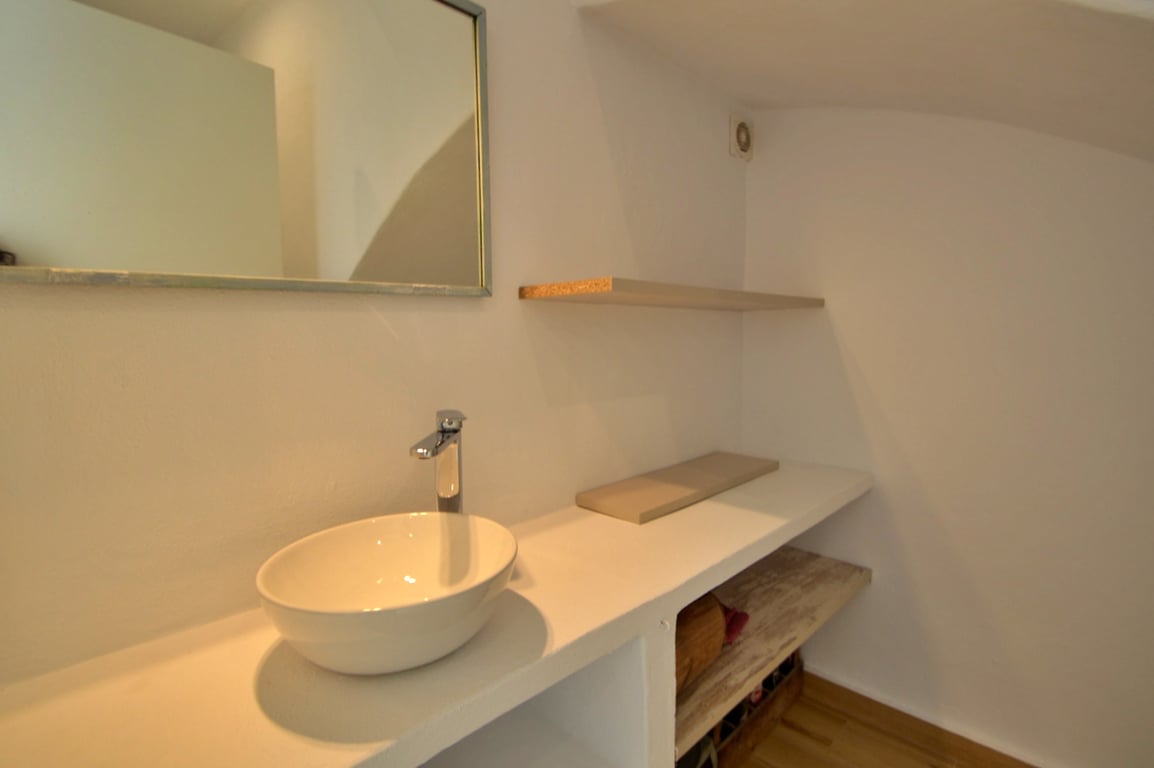 Minimalistische badkamer met een witte opzetwastafel, ingebouwde open schappen en een rechthoekige spiegel. De lichte muren en houten details zorgen voor een strakke afwerking.