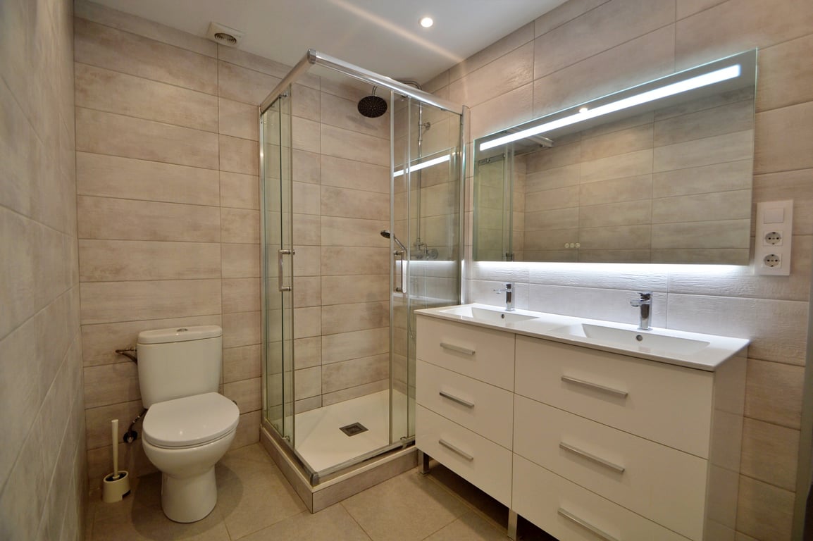 Moderne badkamer met dubbele wastafel, wit meubel, verlichte spiegel en een glazen hoekdouchewand met beige wandtegels.