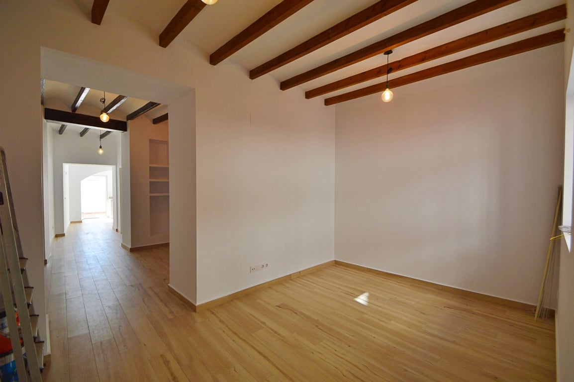 Open ruimte met authentieke houten plafondbalken, lichte vloeren en witte wanden. De hangverlichting en natuurlijke lichtinval benadrukken het moderne, gerenoveerde karakter.