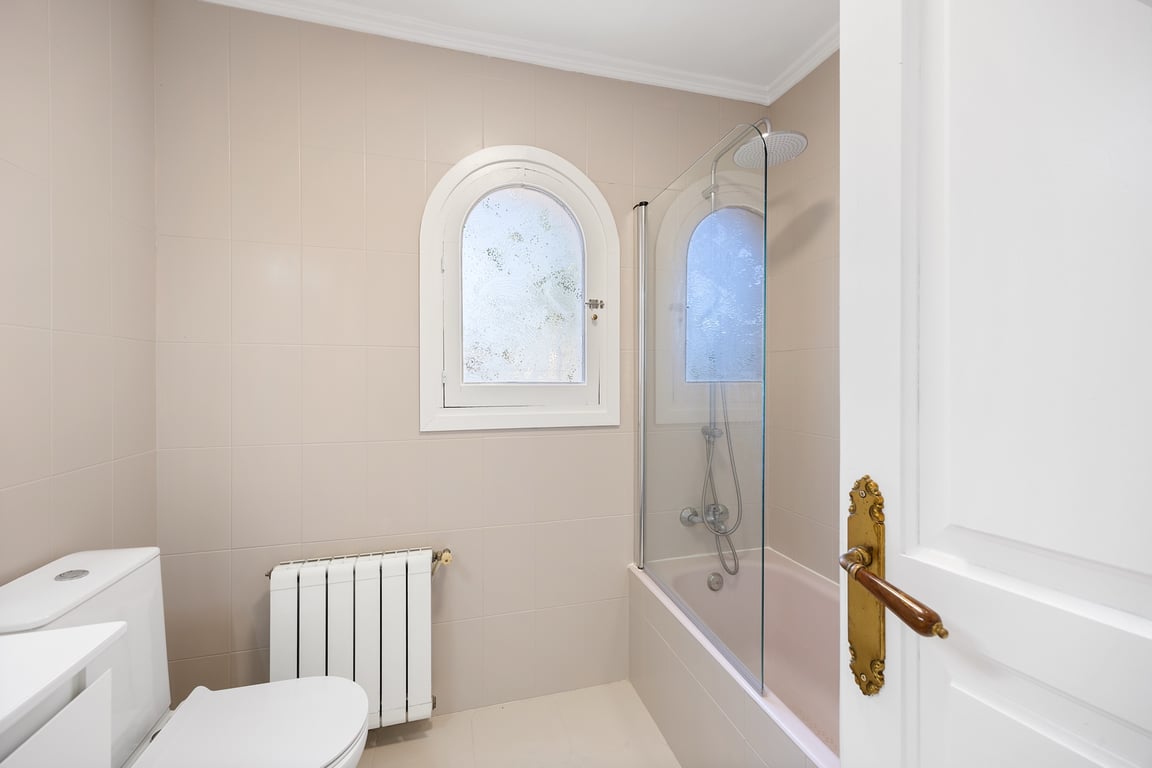 Functionele badkamer met ligbad, glazen douchescherm en een karakteristiek rondboogvenster. Voorzien van radiatorverwarming en neutrale wandtegels.
