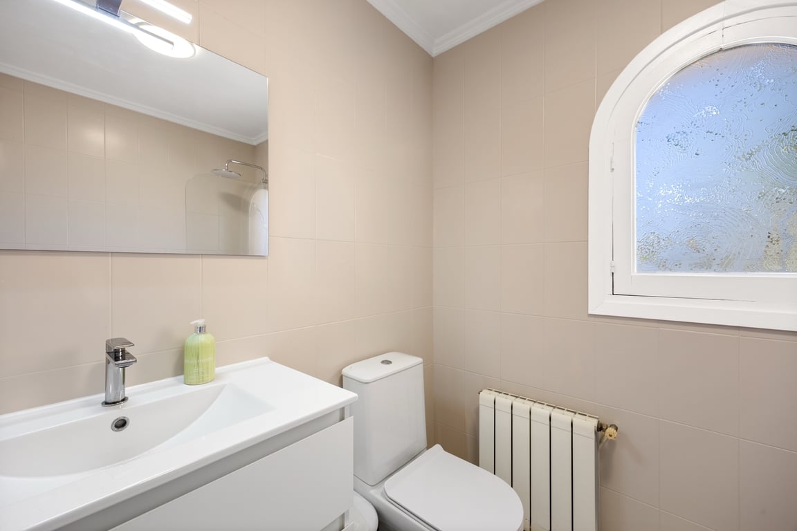 Functionele badkamer met neutrale betegeling, wit wastafelmeubel, radiator en een karakteristiek boograam met matglas voor natuurlijke lichtinval.