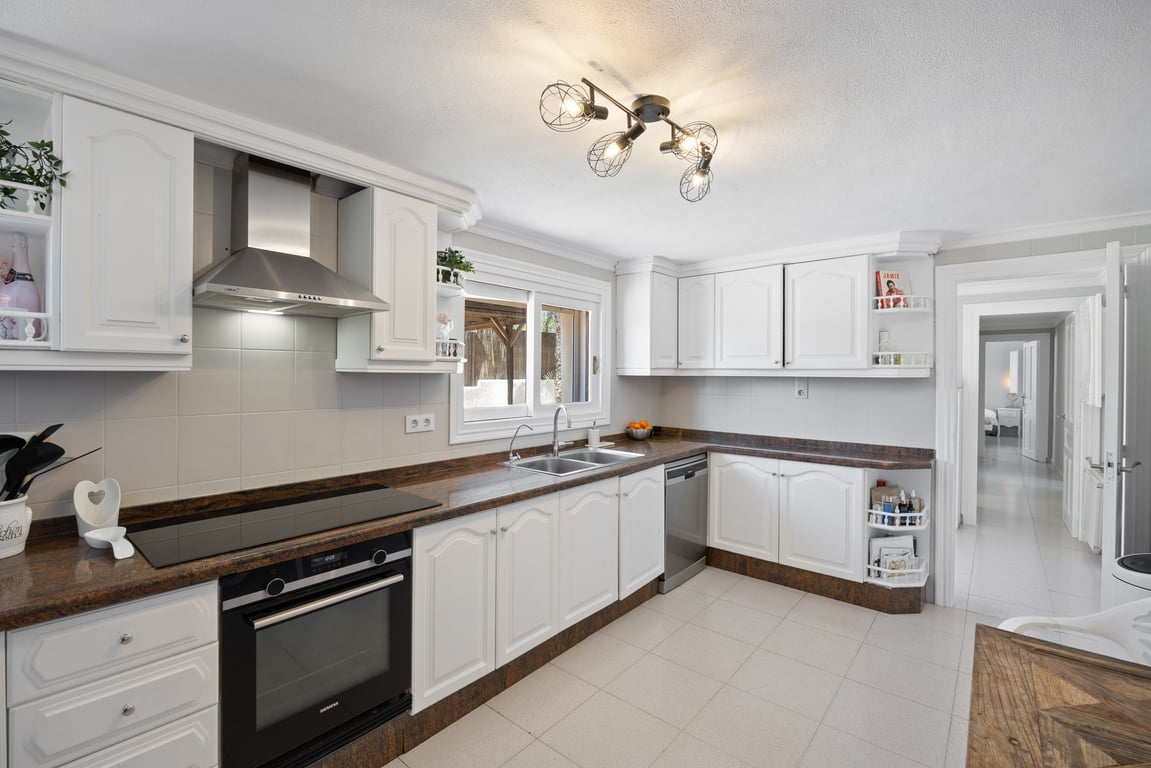 Functionele keuken met traditionele witte kasten, een donker granieten werkblad en RVS-apparatuur. Voorzien van een raam boven de gootsteen en een moderne industriële plafondlamp.
